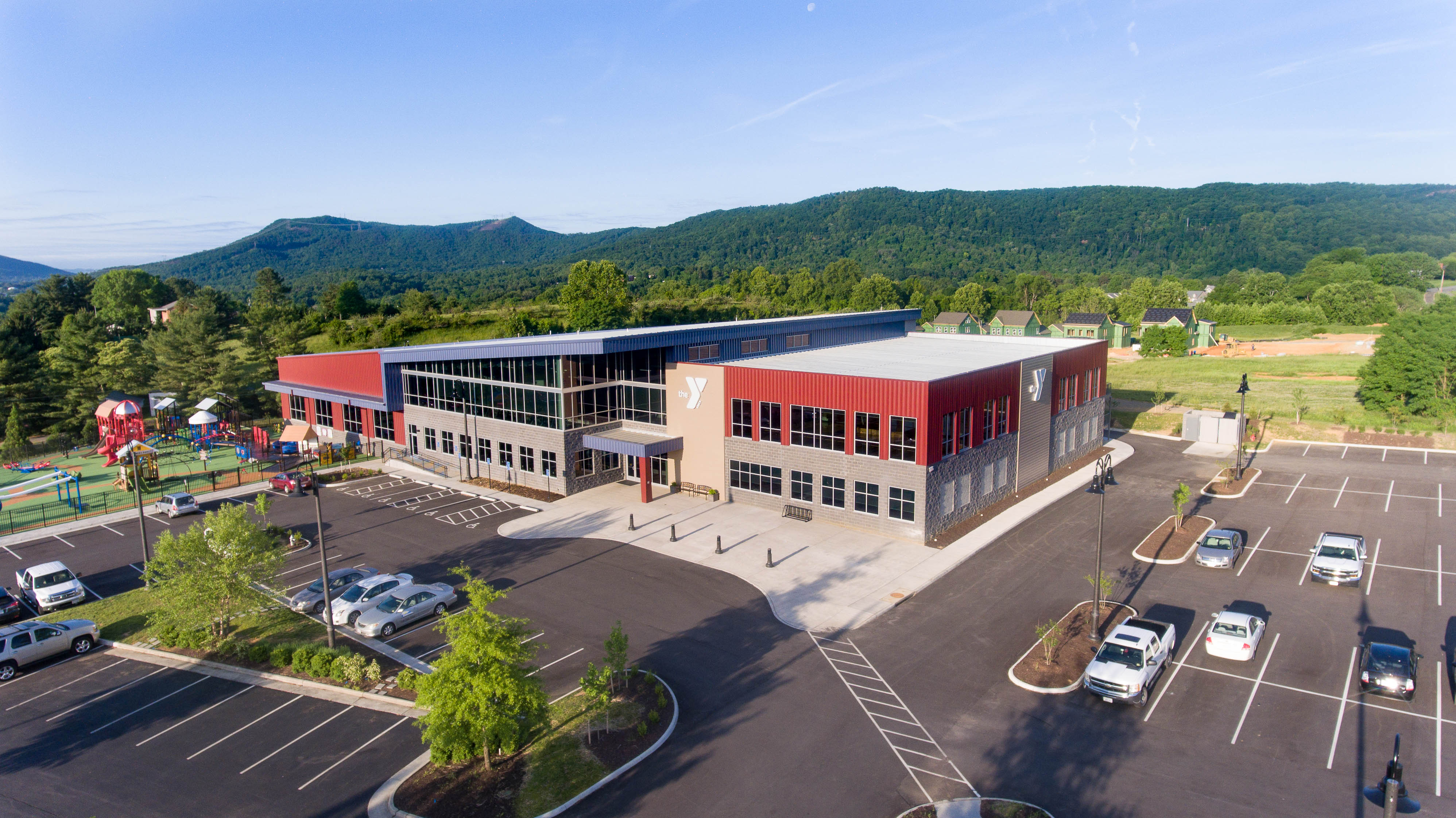 Aerial view of the Botetourt Family YMCA facility surrounded by mountains, with modern architecture, large parking area, and an adjacent outdoor playground under a clear blue sky.