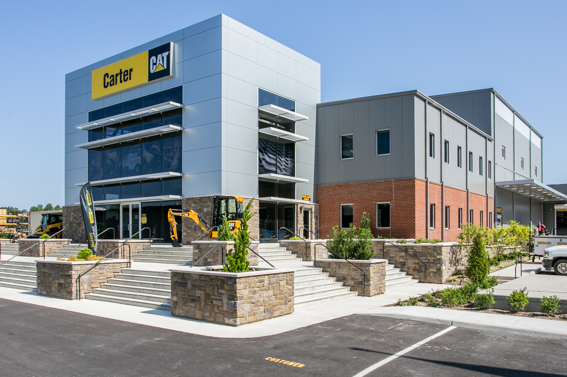 Exterior of the Carter Machinery facility featuring a modern metal and glass facade with a yellow CAT sign, flanked by stone steps, landscaping, and compact construction equipment at the entrance.