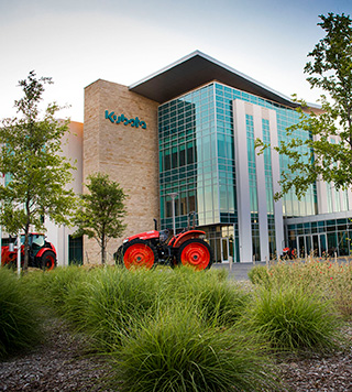 Image of Kubota Headquarters. Several trees and bushes out front of the building and an tractor.