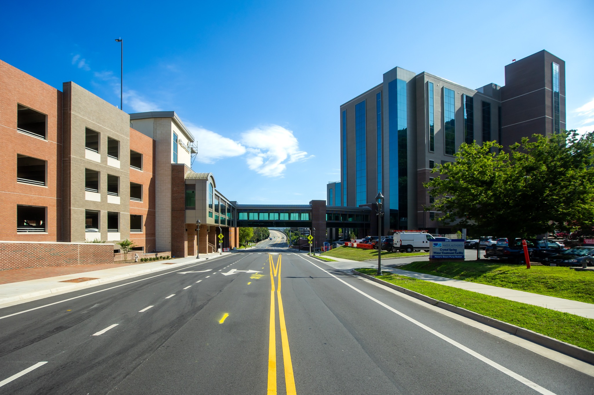 Wide view of a hospital corridor with modern glass pedestrian bridges connecting a parking structure and medical office building, under a bright blue sky on a clear day.